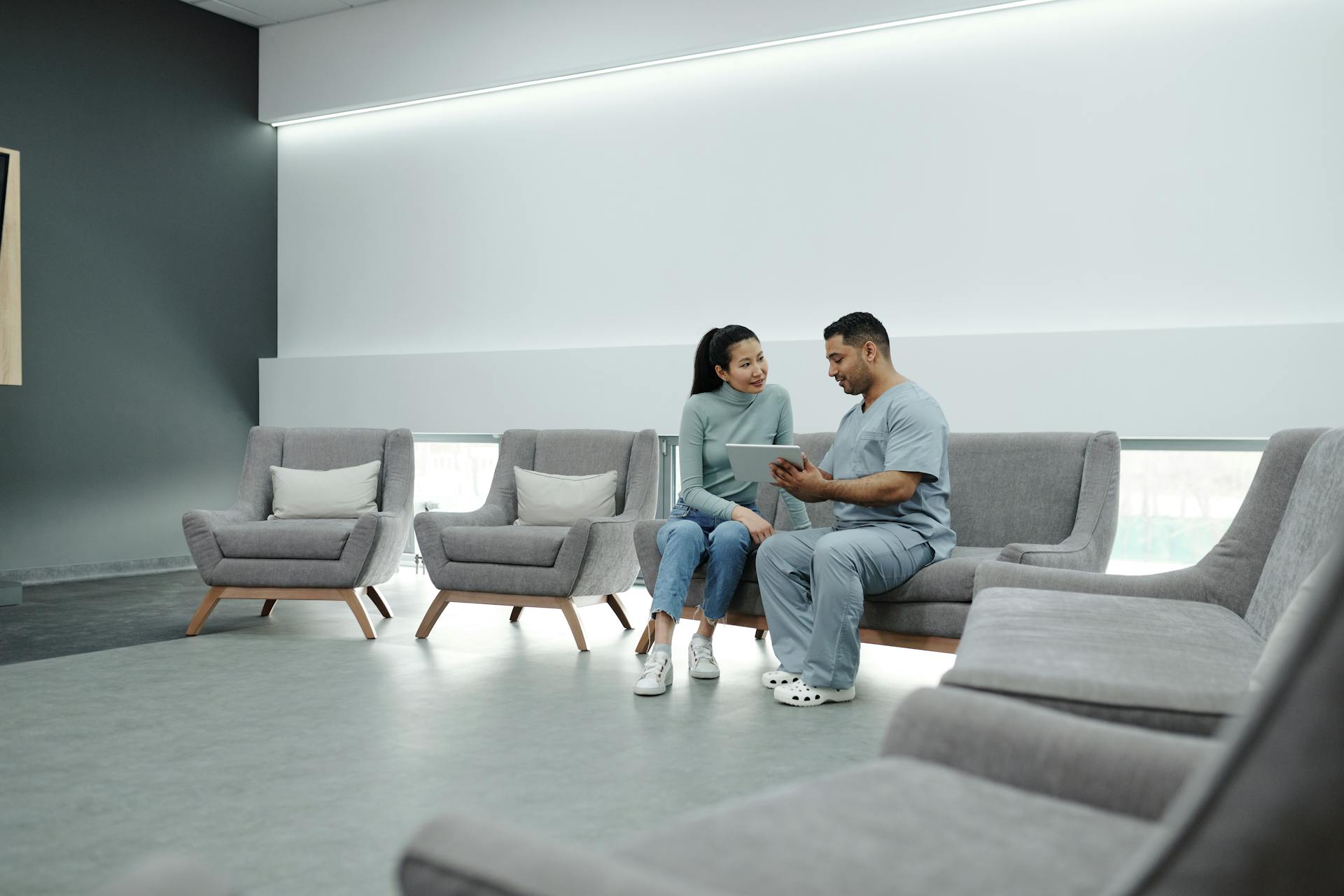 A doctor in scrubs consulting a female patient in a modern clinic waiting room, using a tablet.