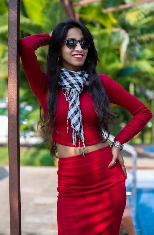 Cheerful graceful ethnic female wearing trendy outfit and scarf standing with hand on waist on sunny poolside and looking at camera with radiant smile