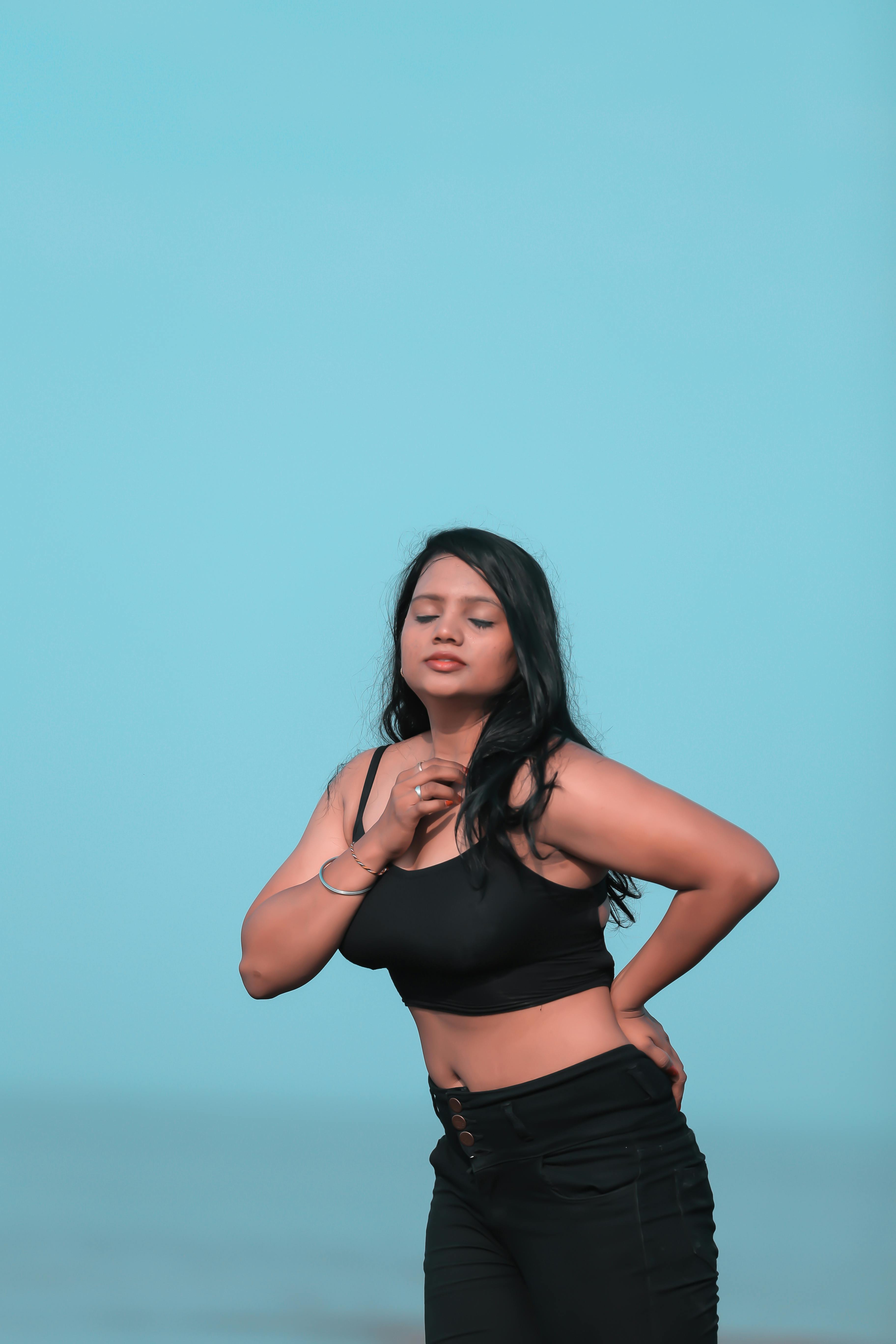 Relaxed ethnic female in black jeans and top standing gracefully with eyes closed and hand on waist against blurred endless sea