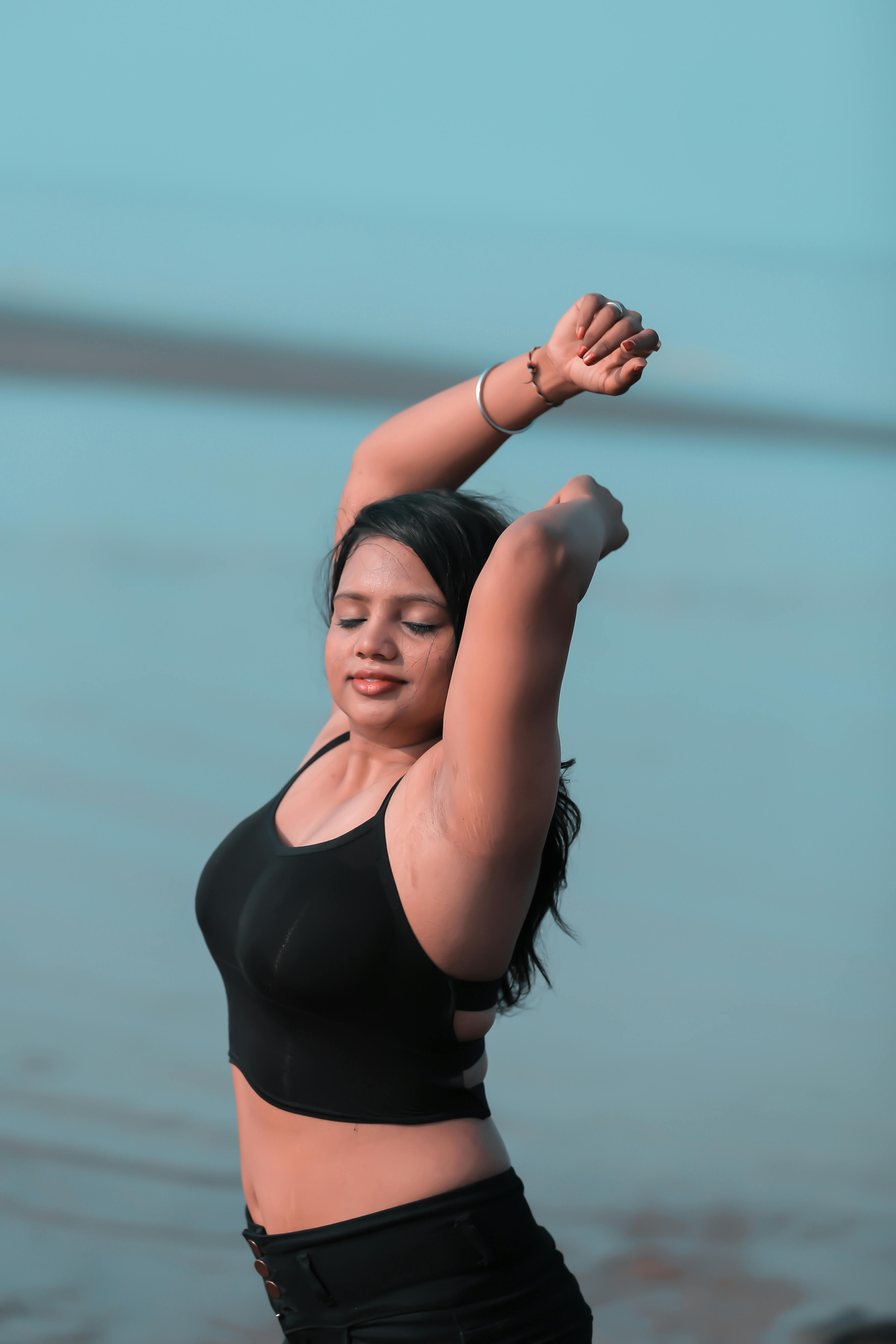 Side view dreamy plump ethnic female in black top with bare belly standing with eyes closed and raising arms gracefully against blurred sea