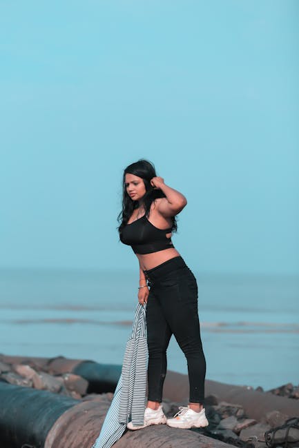 Side view full length contemplative ethnic female in stylish outfit standing on calm stony seacoast while touching hair and looking down with interest