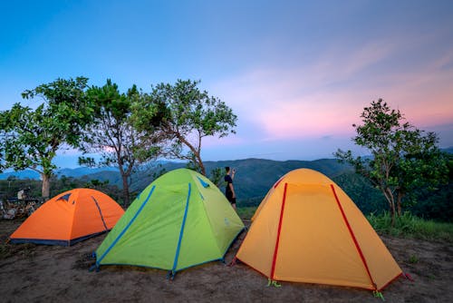 Colorful tents located at camp in mountainous valley near green trees against picturesque sunset sky