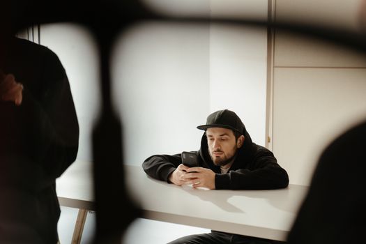 一个年轻人戴着黑色帽子，坐在室内的桌子旁，专注地看着手机