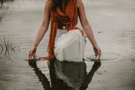 Crop anonymous female in stylish outfit and scarf sitting on haunches in calm pond and touching water