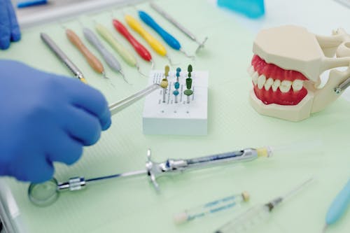 From above of crop faceless orthodontist in latex gloves and tweezers working at medical table with cast jaw and set of syringes near periodontal scalers