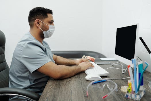 在现代诊所办公室里，一位亚洲男牙医戴着口罩在电脑上打字