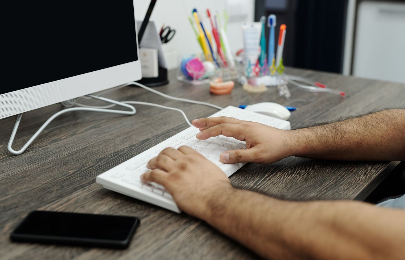 Close-up of hands typing on a white keyboard at a wooden desk in an office setting with accessories.