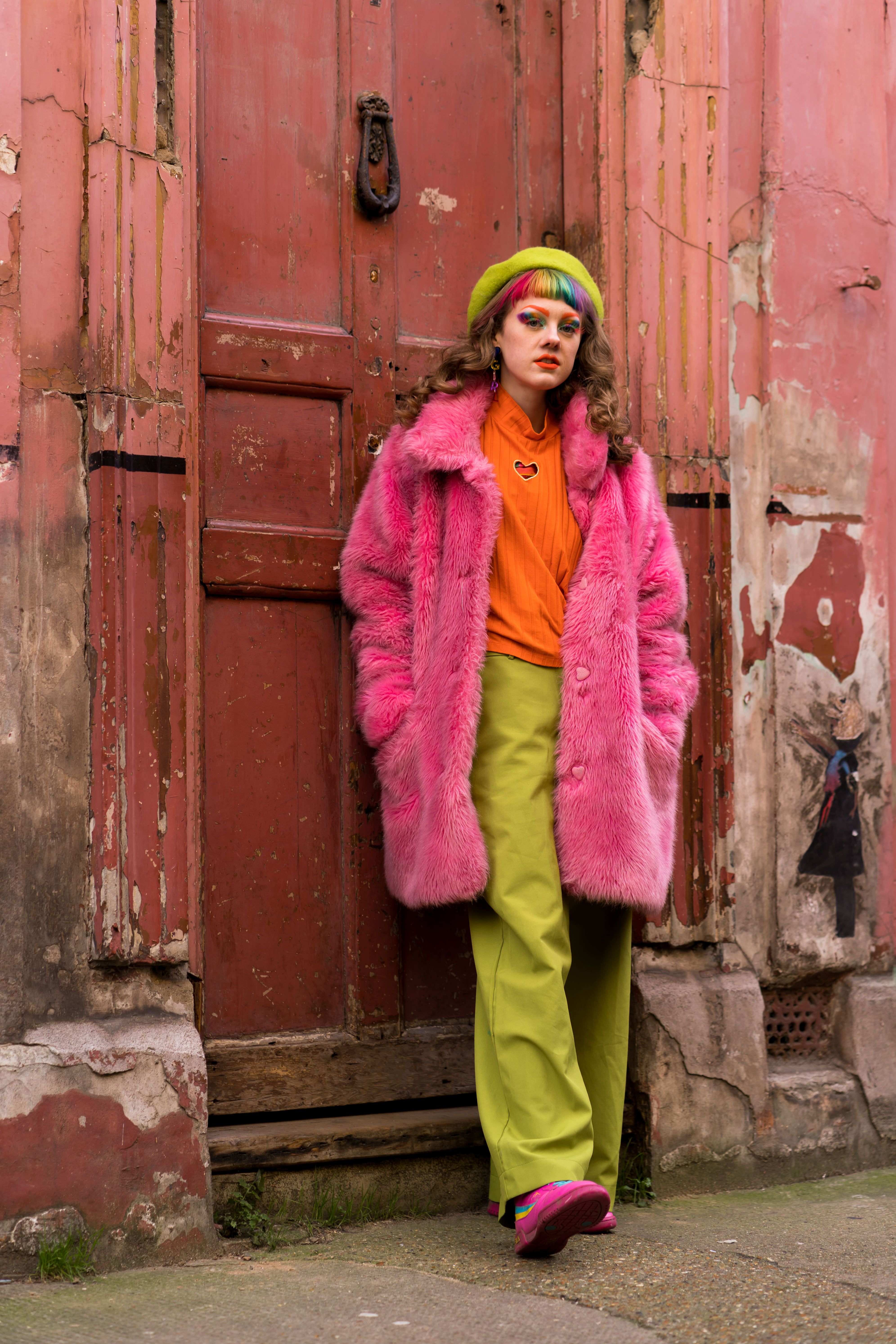 [ColoSach]-full-body-of-young-informal-female-in-colorful-clothes-and-pink-fur-coat-standing-near-door-of-weathered-building-and-looking-at-camera
