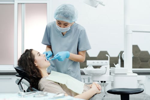 Free Woman in Blue Scrub Suit Standing Beside Woman in Brown Shirt Lying on Dental Chair Stock Photo