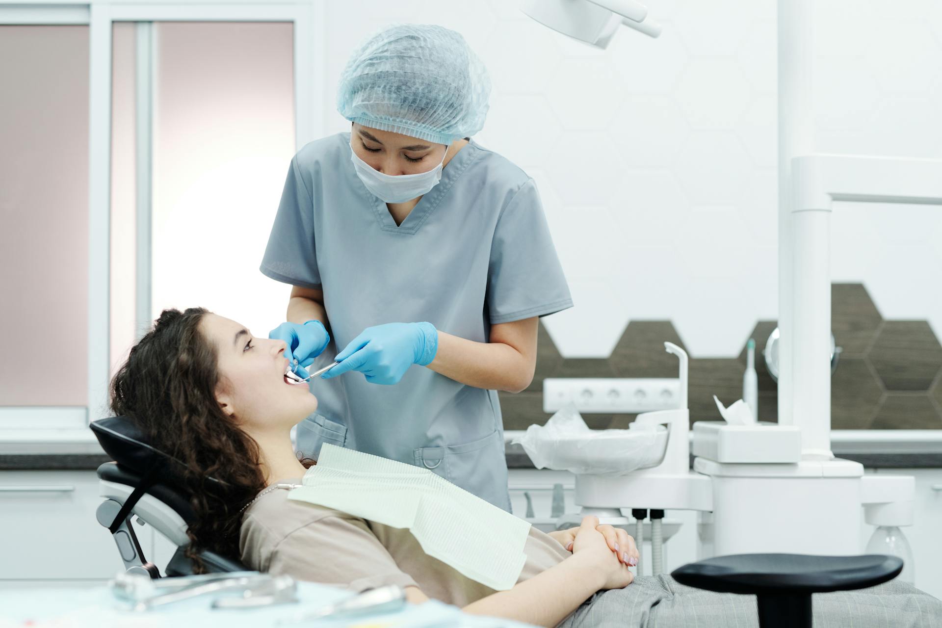 Woman in Blue Scrub Suit Standing Beside Woman in Brown Shirt Lying on Dental Chair