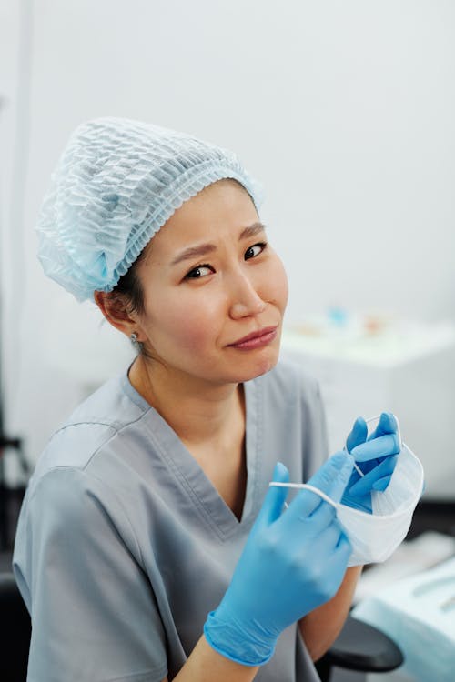 A Woman in Scrub Suit and Latex Gloves Holding a Face Mask