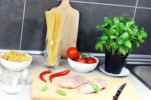 Raw Chicken Breast Seasoned With Peppers Beside Red Chili, Basil, Bowl of Tomatoes, and Raw Pastas on Table