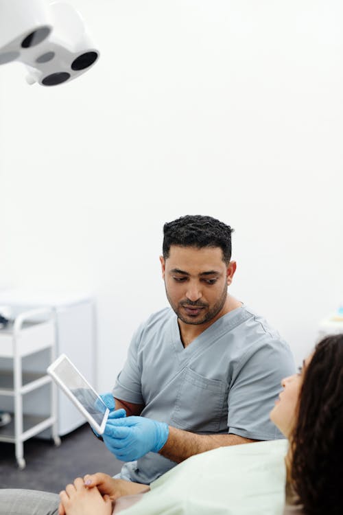 Free Man in Gray Scrub Suit Sitting Beside a Patient while Holding Digital Tablet Stock Photo