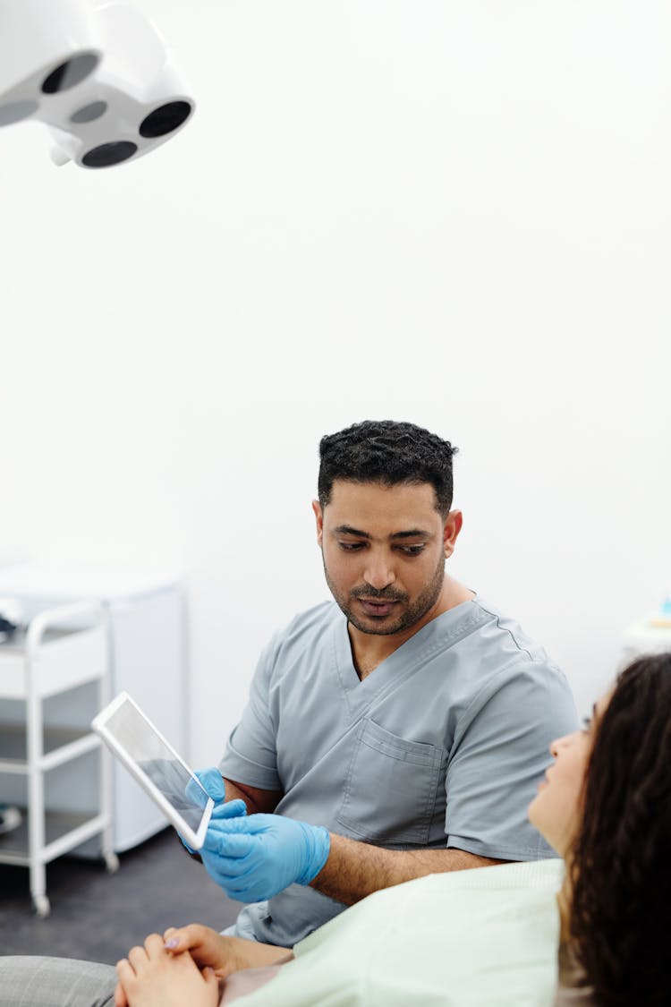 Man In Gray Scrub Suit Sitting Beside A Patient While Holding Digital Tablet