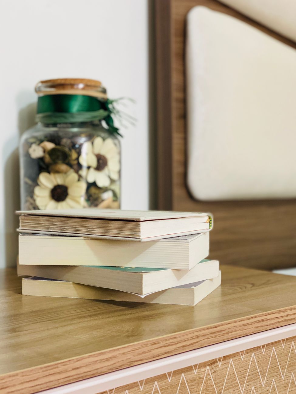 Stack of textbooks placed on wooden table near decorative flower jar in light bedroom