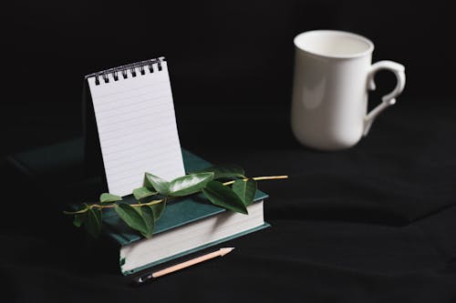 White cup and notepad arranged with book and green leaves on black table