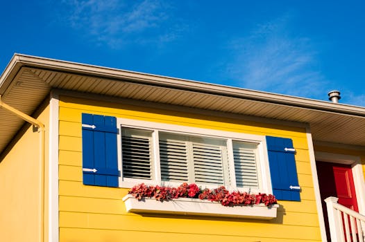 Bright yellow house facade with blue shutters, a window box of flowers, and clear blue sky.
