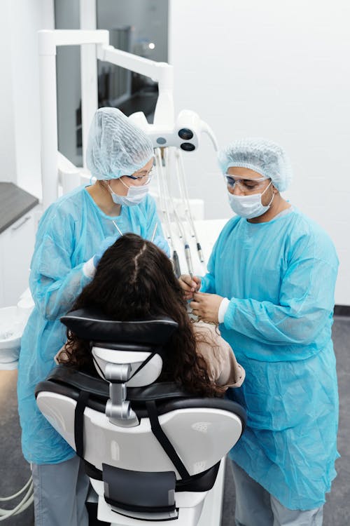 Free Dentists in Blue Scrub Suit Standing Beside the Woman Sitting on the Dental Chair Stock Photo