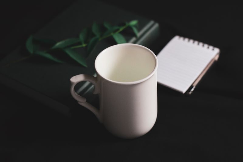 High angle arrangement of white ceramic cup placed on black table near diary and notebook.
