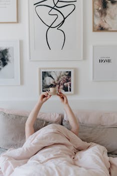 A person enjoys a cozy morning in bed holding a cup of coffee, surrounded by art.