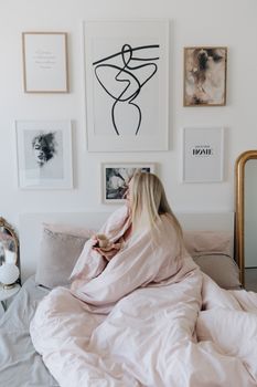 Woman enjoys a cozy morning with coffee in a modern bedroom featuring framed art.