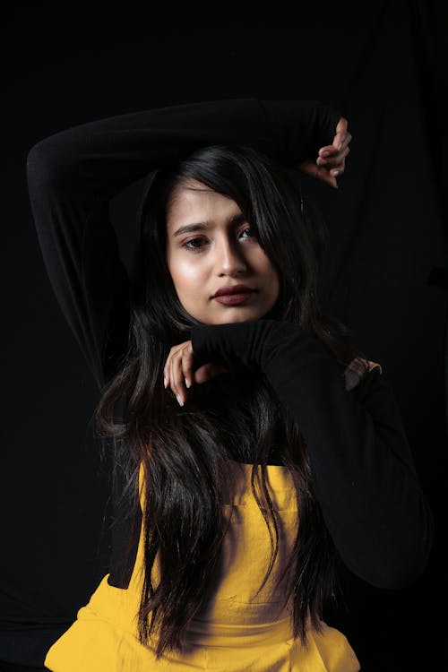 Portrait of unemotional ethnic female in trendy wear raising arms and touching head on black background in modern studio during photo session