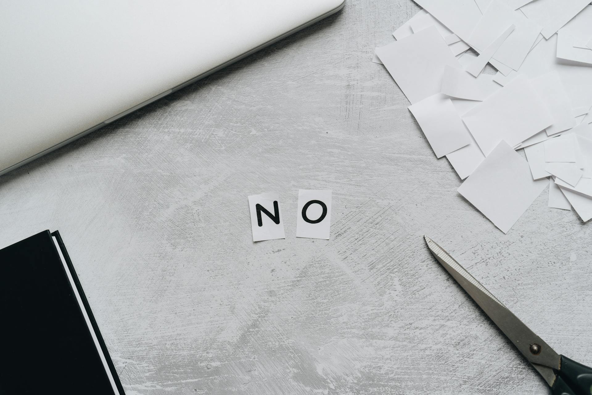 Top view of scattered paper squares, laptop, and scissors forming the word 'NO', implying rejection or denial.