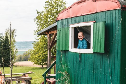 A joyful child smiles from a green camper window set in a scenic countryside.