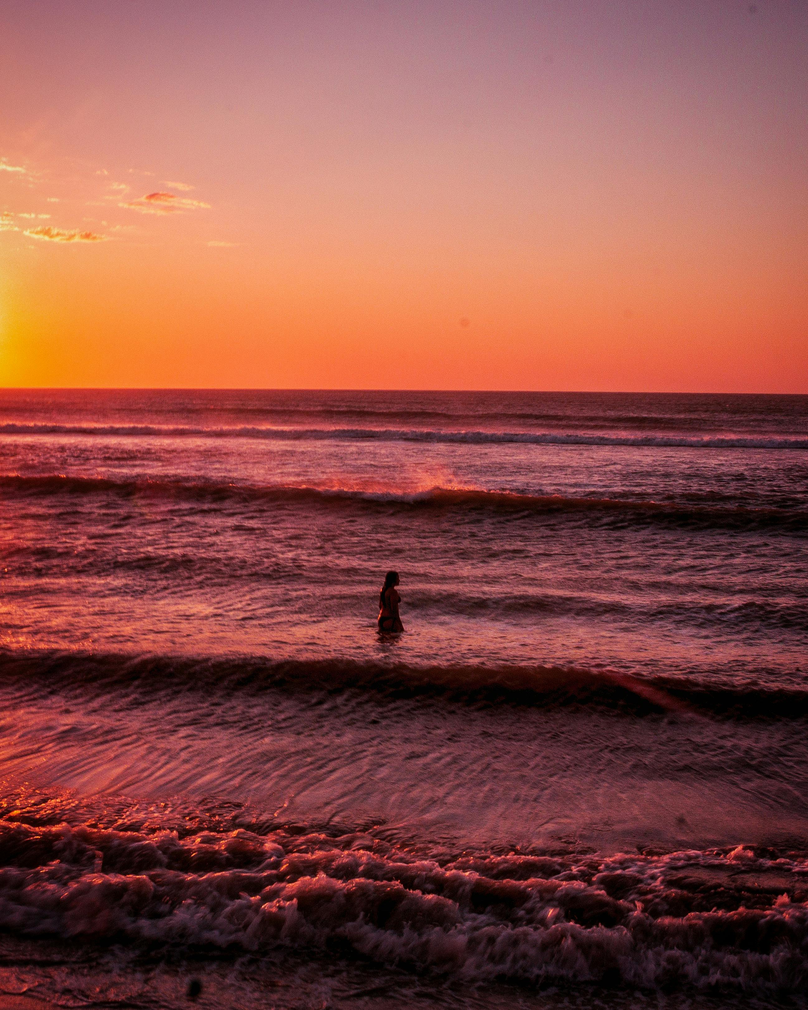 Tranquil scene of a lone person standing in ocean waves during a vibrant sunset.
