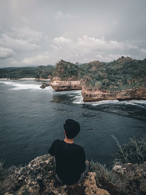 Man in Black Shirt Sitting on Rock Formation Near Body of Water