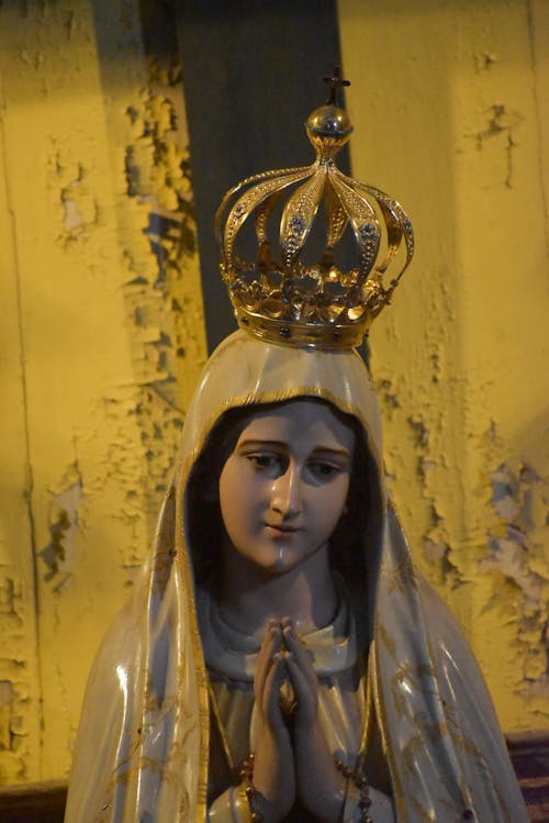 Ceramic figurine of Our Lady of Fatima with hands folded in pray gesture and crown on head against weathered wall