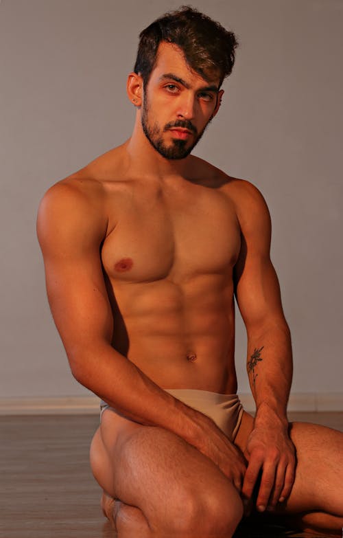 Confident athletic male in skin color underwear sitting on floor in studio