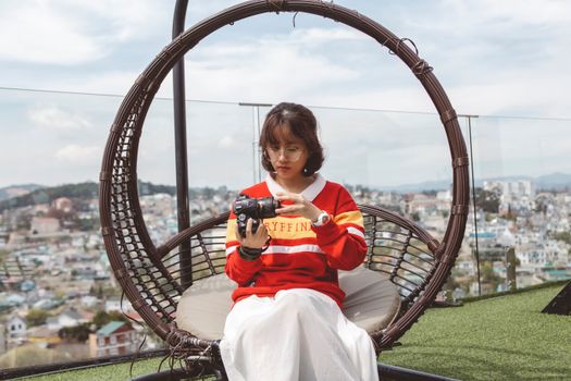 A woman in a red sweater examines a DSLR camera while sitting on a rooftop terrace.