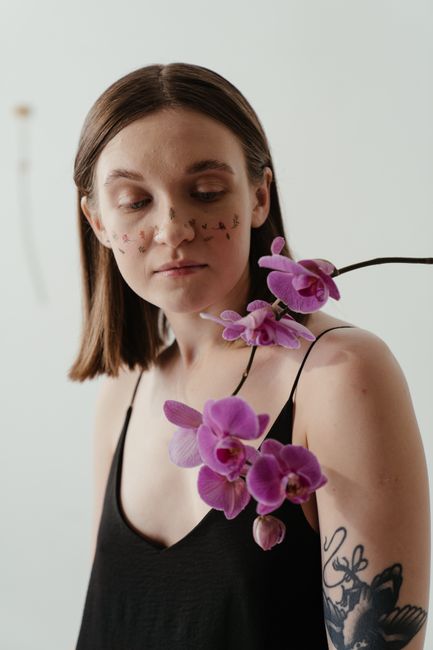 A woman with floral temporary tattoos poses with vibrant purple orchids indoors.