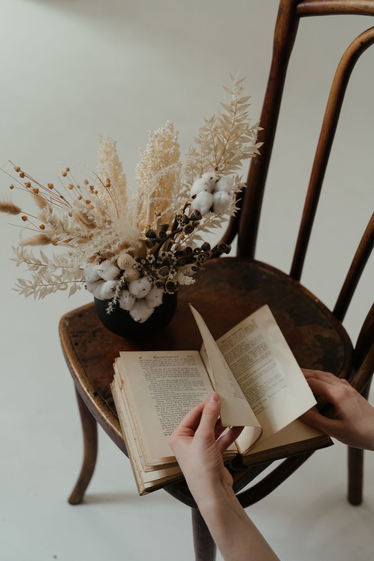 Hands holding an open book beside dried flowers on a chair