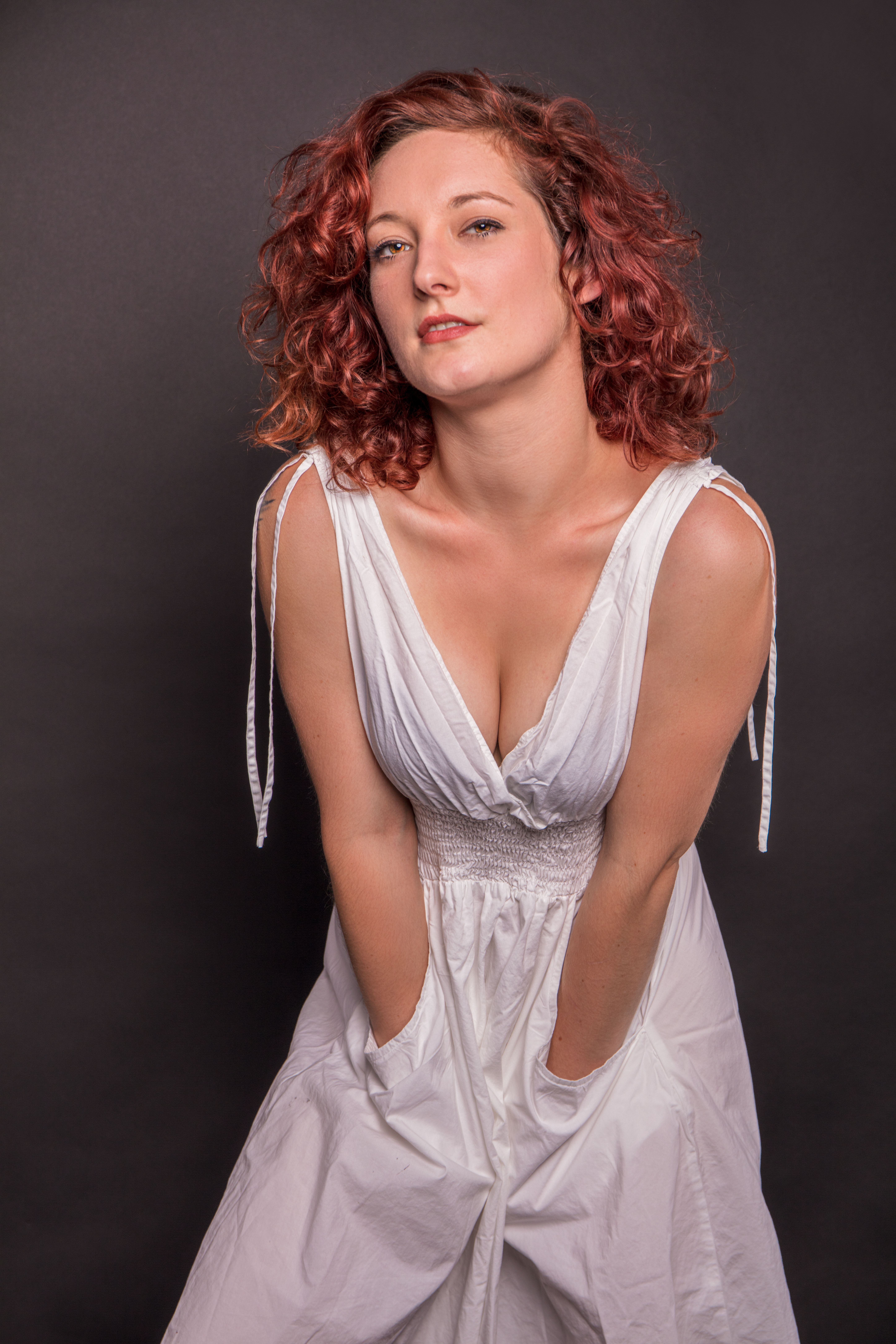 Portrait of a stylish redhead woman in a white dress posing confidently in a studio setting.