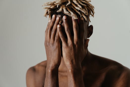 A shirtless young man with dreadlocks covers his face in a contemplative pose, expressing emotion.