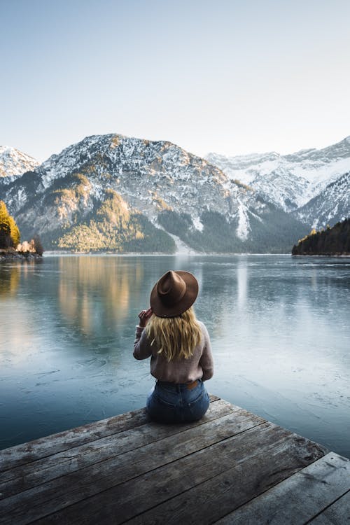 Woman in Brown Jacket Sitting on Wooden Dock Near Lake