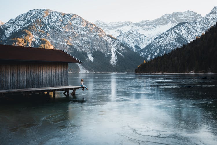 A Person Sitting On A Dock Near The Frozen Lake