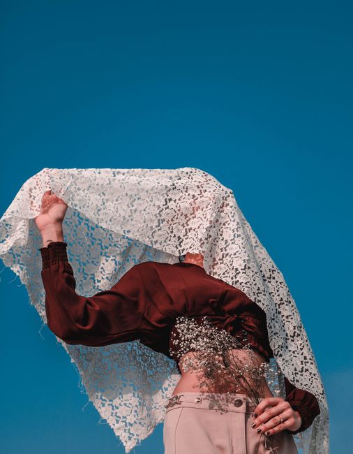 From below woman in elegant blouse holding blossom branch covering head with lace veil under blue sky