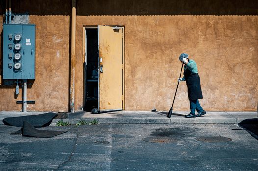 Person sweeping outdoors on a city sidewalk in bright daylight.