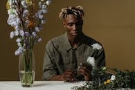 一位留着辫子的年轻人，在宁静的环境中，若有所思地布置着一束花，四周鲜花环绕