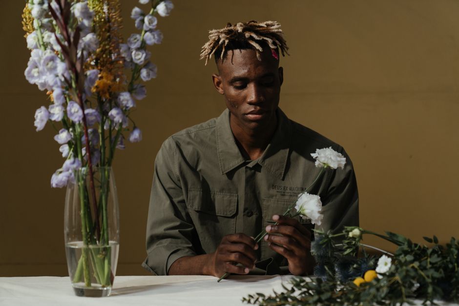 A thoughtful young man with dreadlocks arranges a bouquet, surrounded by blooms in a serene setting.