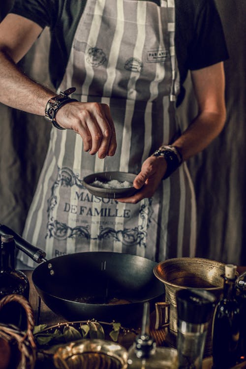 Faceless man in apron taking pinch of salt from bowl while preparing hot dish in metal wok pan