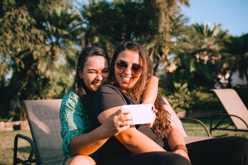 Free Cheerful woman embracing female friend in sunglasses while sitting together on lounger and taking selfie on cellphone near growing trees under blue sky in summer on weekend Stock Photo