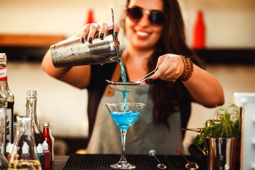 Free Crop smiling female in sunglasses pouring bright alcoholic drink from shaker into glass using small sieve while standing at bar counter Stock Photo