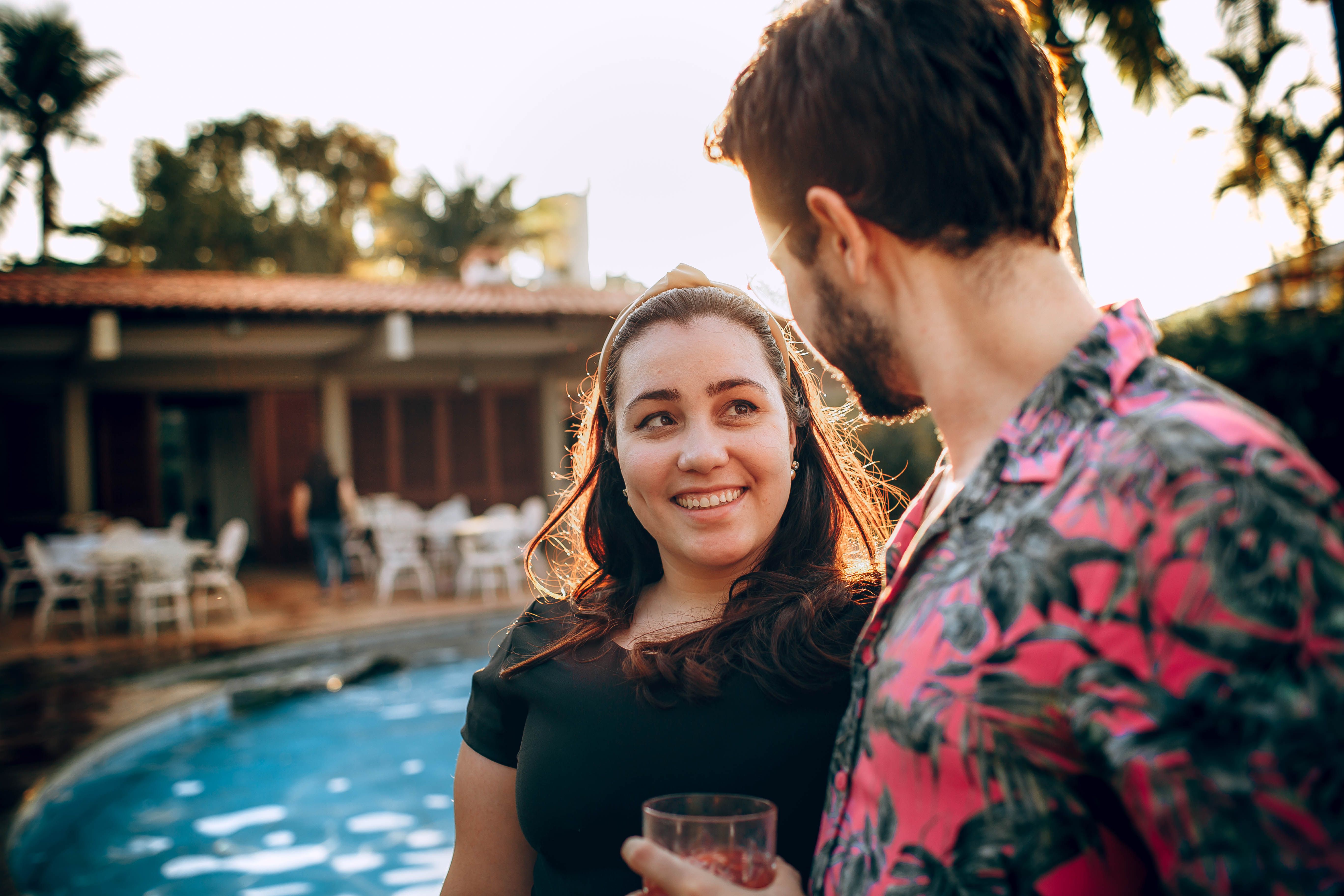 Content woman conversing with crop unrecognizable boyfriend while standing together near swimming pool and trees under sky during honeymoon in back lit and looking at each other