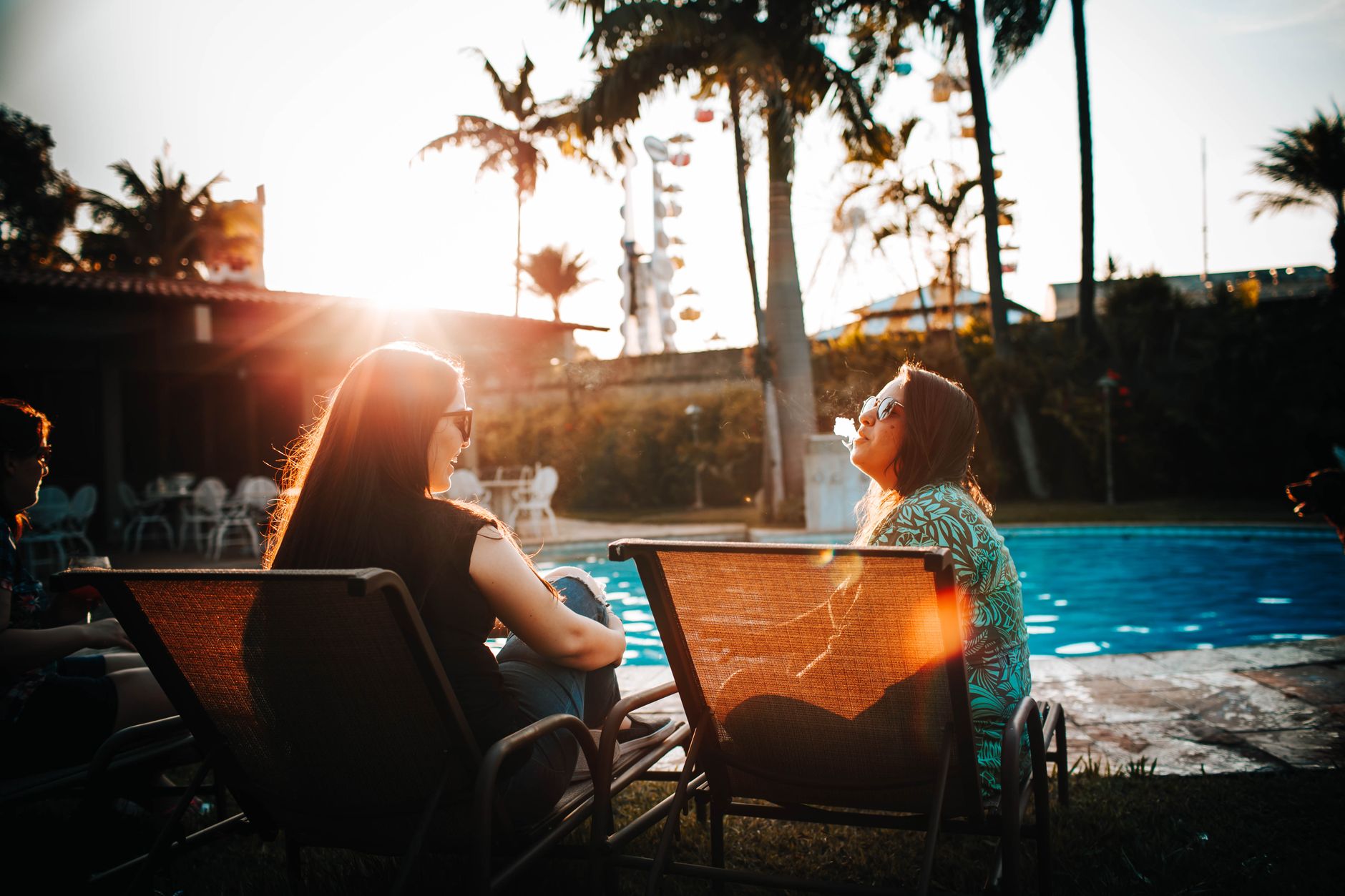 Two women sitting on lounge chairs by a pool, enjoying a sunny day outdoors.