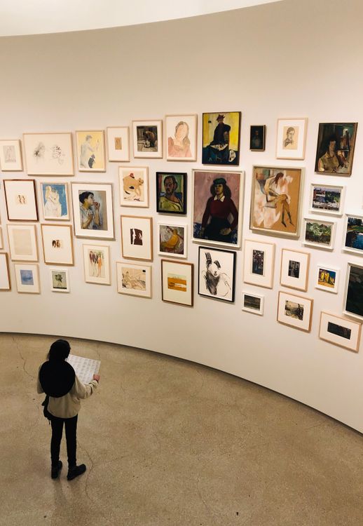 Free From above back view of anonymous female with paper sheet standing on floor in corridor in front of wall with pictures in frames at exhibition in art gallery Stock Photo