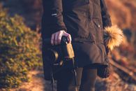 Side view of crop anonymous woman in outerwear holding professional contemporary photo camera while standing with knitted hat with pompon near green bush in daylight